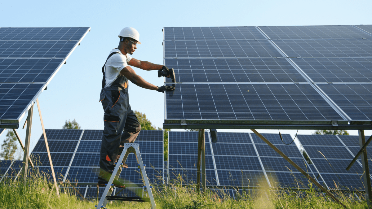 A person in a white hard hat stands between two solar panels, working on the one to the right