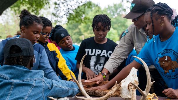 Thank-a-Ranger Students gather around a table outside surrounded by trees with outstretched hands to touch a variety of pelts and bones as part of an environmental education program led by a park ranger at a local park