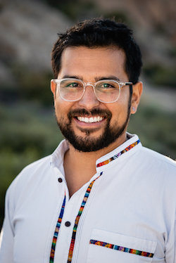 Photo of Ángel Peña who has short black hair and wears glasses and a white tunic with colorful embroidery. 
