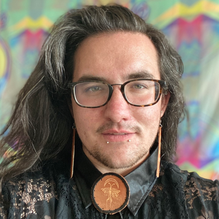 Photo of Grant Gliniecki who wears round glasses, has long wavy brown hair, and tan leather jewelry.