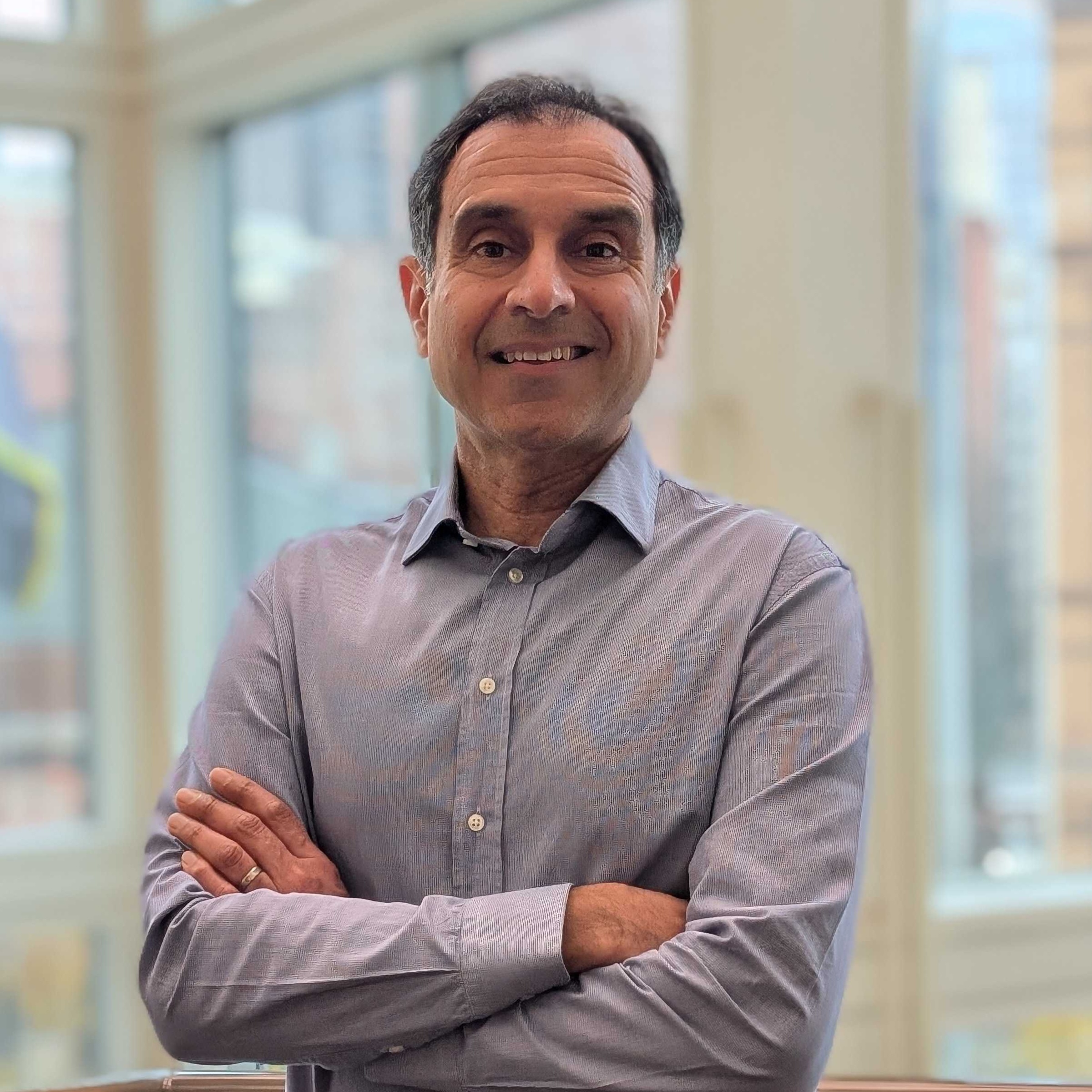 Vik Mohan stands in front of a large window, smiling with his arms folded