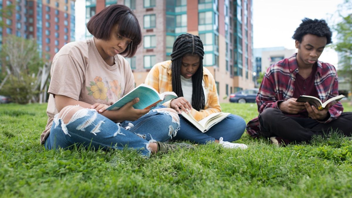 Three persons reading books while sitting on grass outside