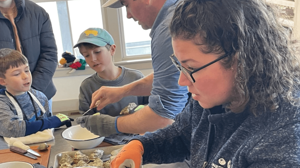 A curly-haired woman wearing glasses and orange gloves holds an oyster. In the background are two young boys watching as an adult shucks an oyster. They are all indoors.
