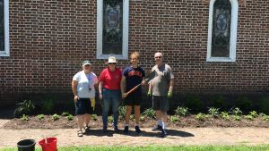 A group of four people stand, facing the camera, in front of a brick building with a garden bed.