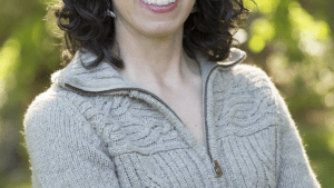 image of Jeanine Silversmith with medium length dark curly hair, glasses, smiling, and wearing a light gray sweater, standing in front of leafy green trees