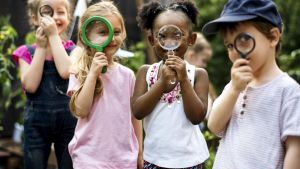 four young children holding magnifying glasses outside in green space
