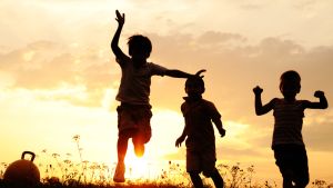 three children running and jumping and backlit by setting sun, peach and yellow colored sky