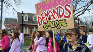 Dozens of teenage students walk through the street. One holds a sign saying "Progress demands change."