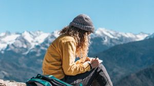 person with long curly hair wearing grey knit cap, yellow top, grey pants, red hiking shoes, sits on mountain top writing, blue backpack sits behind them