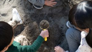 Three kids in a sandbox where one of the kids holds a scooper filled with sand