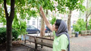 A person in a gray head scarf and green t-shirt reaches up to touch a green leaf on a tree in a city park