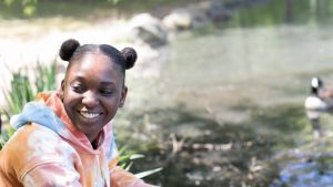 A smiling Black woman looks off to the side while near a duck pond