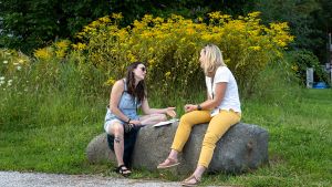 Two people sit and talk with each other on a large boulder outside. In the background, a stand of tall yellow flowers.