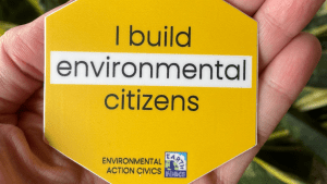 A hand holds a yellow pin that says "I build environmental citizens." Earth Force logo is at the bottom.