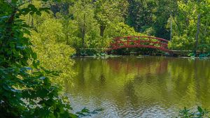 A small lake surrounded by forest in summer foliage. In the distance, a red arched bridge extends over a small portion of the lake. Above, blue sky.