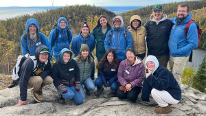 A group of Wolf Ridge naturalists stand atop a rocky outcrop overlooking forested bluffs and Lake Superior.