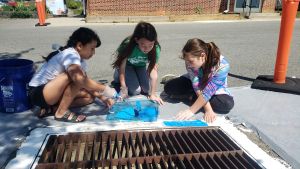 Three kids sit next to a storm drain along a road with a brick building in the background. The storm drain has white paint bordering the drain. The kids have a stencil on top of the white paint, and are painting blue on top of the stencil to create messaging around the storm drain.