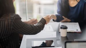 Two people seated across from each other at a dark wood table shake hands; beneath them on the table is a clipboard, papers, a coffee cup, and a smartphone