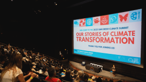 Photo of an audience sitting in an auditorium and watching a large screen of a slide that says "Our Stories of Climate Transformation"