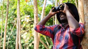 Smiling person in red flannel and fishing hat looking through pair of binoculars in wooded area