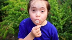 A child outside blowing dandelion seeds in front of greenery