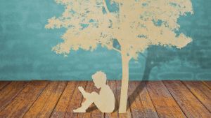 white paper cutout of tree and boy sitting under it reading while sitting on hardwood floor with painted blue background