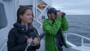 Two researchers looking at the ocean from a boat. One has a binocular while the other is taking notes.
