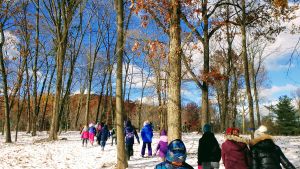 A group of nearly 20 children, bundled up in winter attire, walk into a snowy landscape with sparse trees. In the background, a dense forest in winter, and blue sky with light clouds above.
