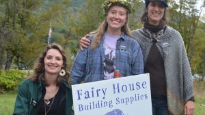 Three light-skinned and long-haired people smile behind a yard sign that reads "Fair House Building Supplies." The person to the left crouches, and the two people to the right stand. Behind them, green grass and trees in late summer foliage.