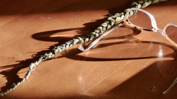 A strand of braided sweetgrass and a pair of glasses rest on a table with the sun created shadows across them. Photo credit:Judith Jackson