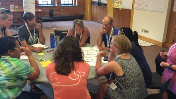 Group of adults with thoughtful expressions gathered around a round table with markers and chart paper. 