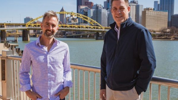 Ryan Rydzewski and Gregg Behr stand in front of a yellow bridge in Pittsburgh, Pennsylvaina.