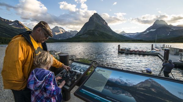 An adult an a child read an interpretive sign next to a mountain range and a body of water