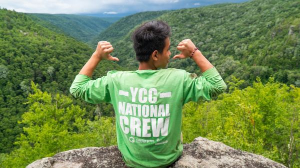A young person sitting on the edge of a boulder overlooking forest-covered hills. The person faces away from the camera, while pointing at their green shirt that says, "YCC National Crew."