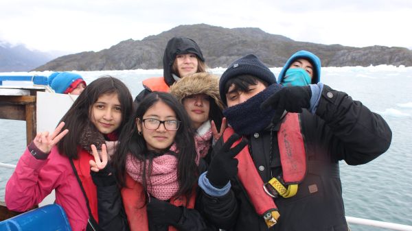 six youth bundled in warm clothing stand before a body of water with a mountain in background