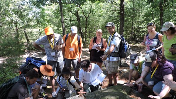 A group of people huddled in a forest with open notebooks. Some are kneeling examining a rock's surface or the ground.