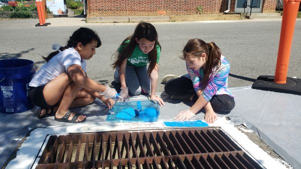 Three kids sit next to a storm drain along a road with a brick building in the background. The storm drain has white paint bordering the drain. The kids have a stencil on top of the white paint, and are painting blue on top of the stencil to create messaging around the storm drain.