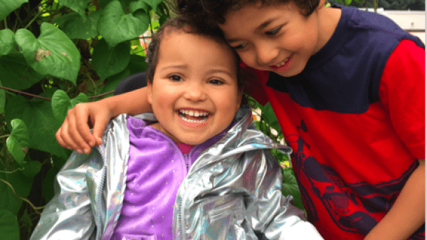 Two kids hug, one use a wheelchair they are in front of a green nature filled background with the text of the book title on top. 