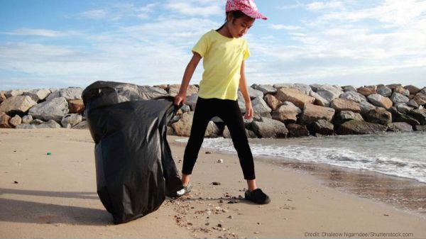 A child looks down at a sandy beach while holding a trash bag