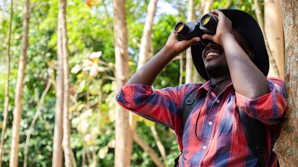Smiling person in red flannel and fishing hat looking through pair of binoculars in wooded area