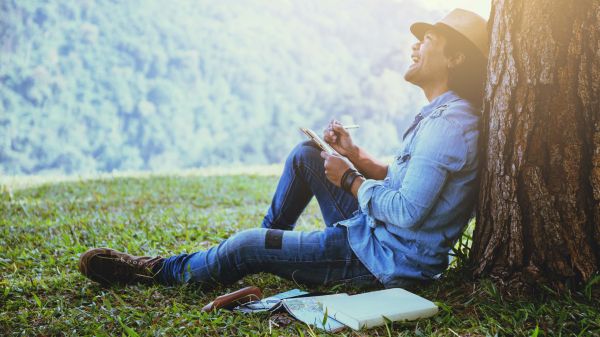 man sitting against tree writing in notebook and looking up at trees on hillside