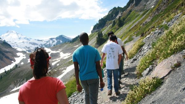 A line of four teens hiking on a mountain trail. Snow-capped mountains in the distance.
