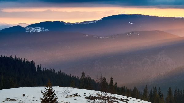 Photo of a snowy hill, forest silhouette in the back, and rows of mountain ranges higlighted by a few rays of sunlight peeking out under a dark blue cloudy sky