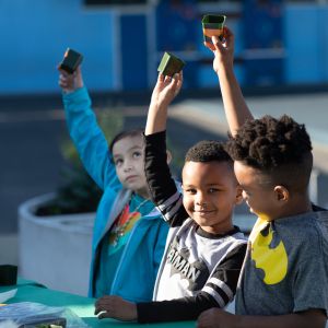 Three young kids hold up stamps. The child in the middle smiles as the other child.