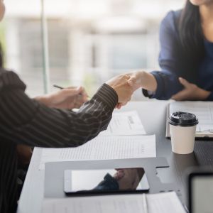 Two people seated across from each other at a dark wood table shake hands; beneath them on the table is a clipboard, papers, a coffee cup, and a smartphone