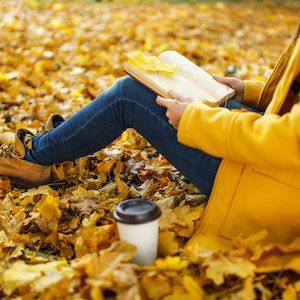 Person sitting amongst fallen yellow and orange leaves, wearing yellow raincoat and reading a book.
