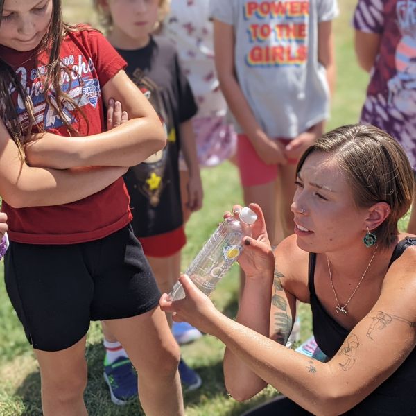 A group of kids huddle around a person kneeling to hold a plastic water bottle out