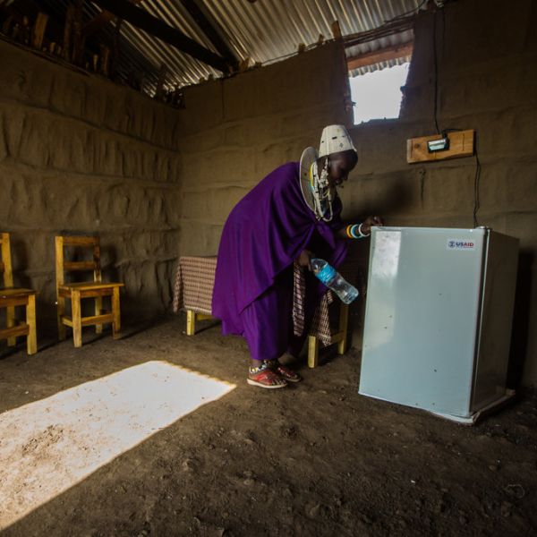 A woman holding a water bottle, stands near a refrigerator in a room