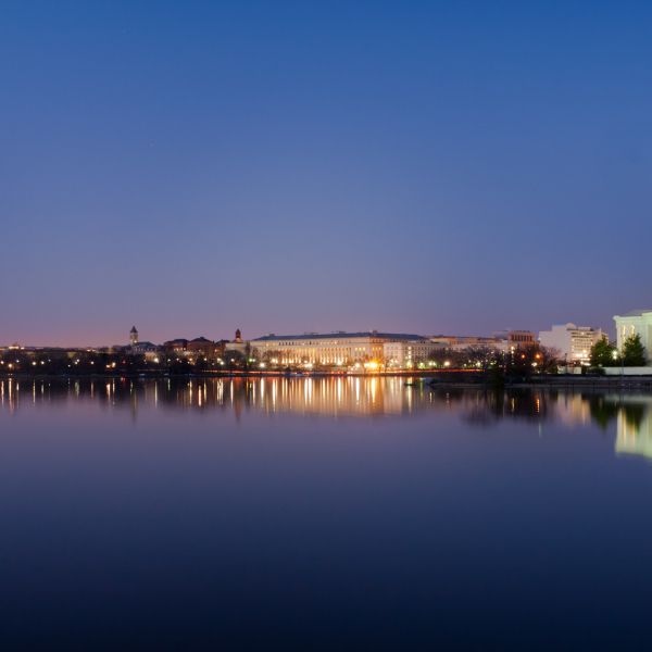 Washington DC at dusk. The Washington Memorial stands illuminated on the left. The Jefferson Memorial, also illuminated stands to the right. The river runs in the foreground. 