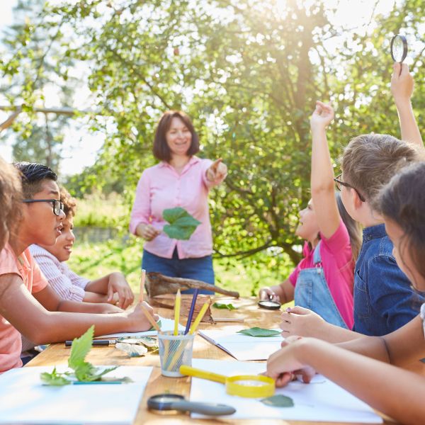 Outdoor classroom, kids at table, adult holding a leaf and pointing to raised hand.